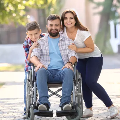 Family smiling on a sidewalk, husband is in a wheelchair 