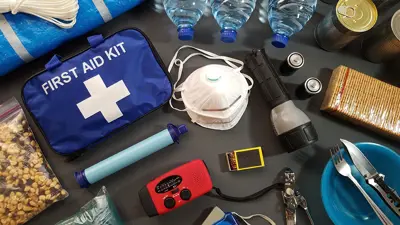 A blue first aid kit and emergency items placed around it, including water bottles, a mask, flashlight, dry food goods, and other tools.