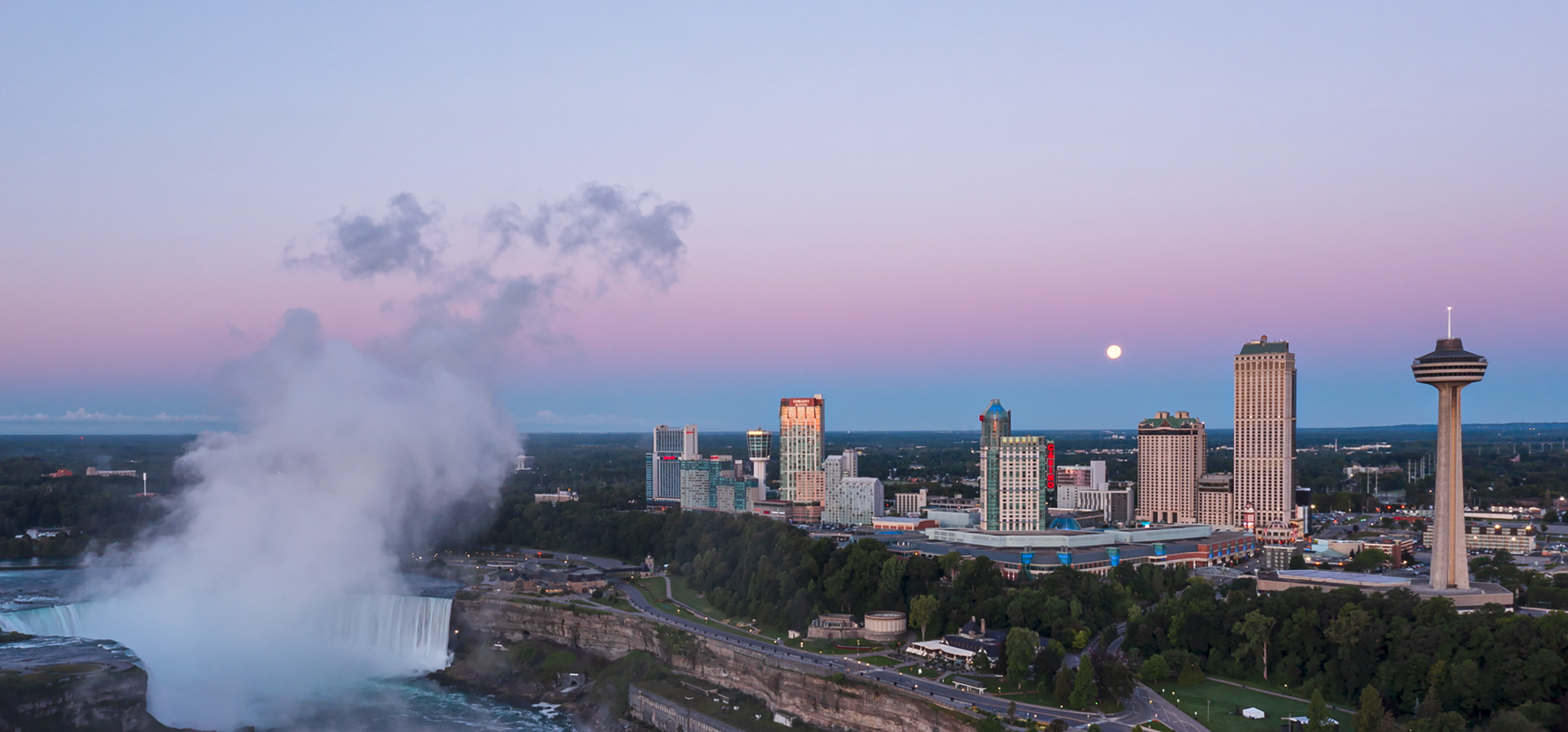Aerial view of Niagara Falls and the mist rising, with buildings in the background.  Showing a sky at dusk with the moon above.