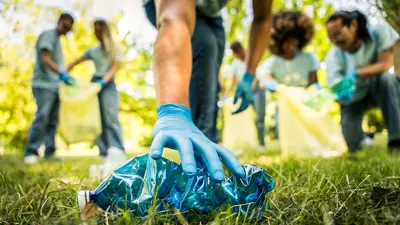 A close up of someone picking up litter off the grass, with volunteers in the background holding garbage bags.