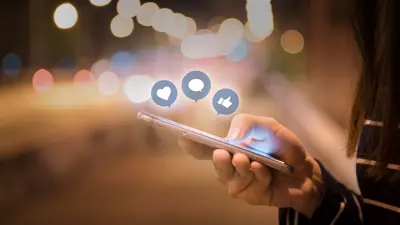 Close up image of a person holding a cell phone and social media graphics coming out of the phone in shape of heart, speech bubble, and thumbs up.