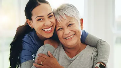 Close up of a woman hugging a senior woman from behind, both are smiling.