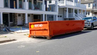 The end of an orange dumpster seen on a street in front of a truck at a construction site on a sunny day.