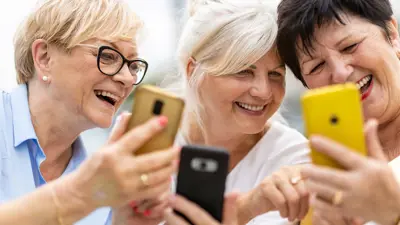 Three senior women with their heads close together, smiling and looking at their phones.